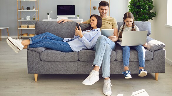 Eine Frau, ein Mann und ein Mädchen sitzt vor elektronischen Geräten auf einem Sofa.