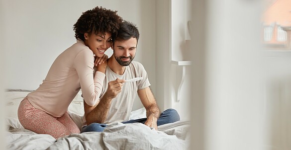Ein junges Paar schaut auf einen Schwangerschaftstest während sie auf einem Bett sitzen.