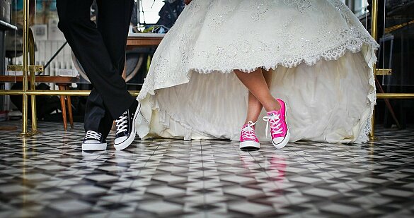 Das Bild zeigt die Beine eines Mannes und die einer Frau, die ein Brautkleid und pinke Schuhe trägt