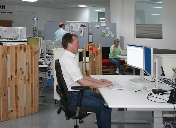 Ein Mann sitzt an einem Schreibtisch vor einem Monitor und im Hintergrund sind weitere zwei Personen zu sehen
