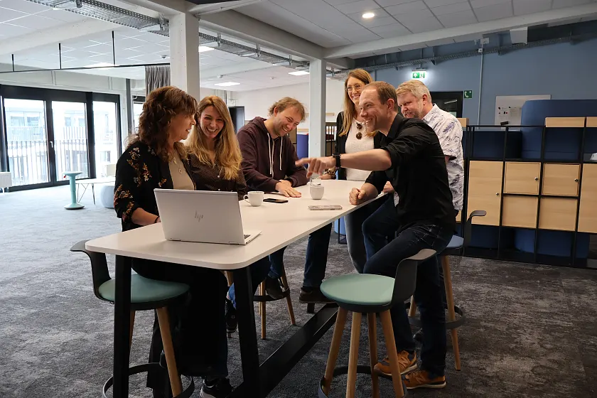 Sechs Personen stehen um einen Tisch und schauen sich eine Pr&auml;sentation auf dem Notebok an.