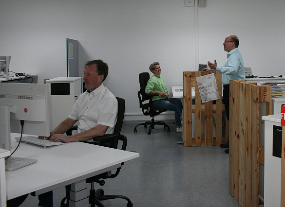 Blick in ein Großraumbüro: ein Mann arbeitet an seinem Schreibtisch, im Hintergrund sind zwei weitere Personen zu sehen