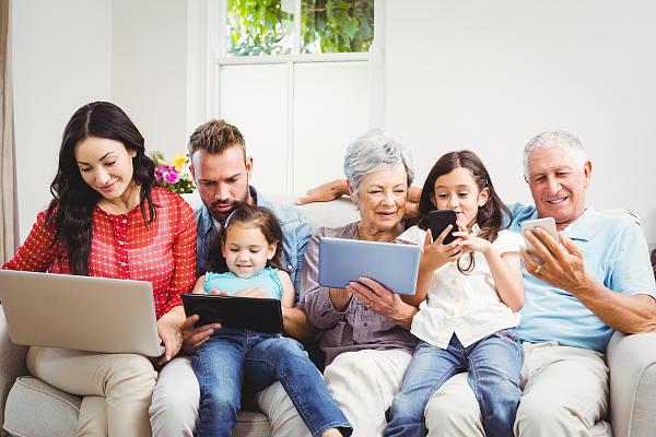 Eine Familie bestehend aus Großeltern, Eltern und zwei Kindern sitzt auf einem Sofa und schaut in Handys oder Notebooks