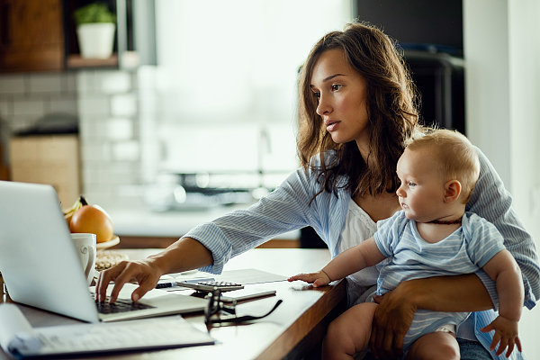 Eine Frau sitzt vor einem Notebook und hält dabei ein Baby auf dem Arm