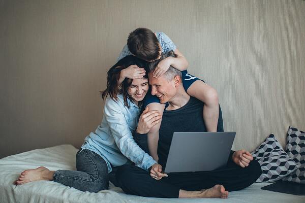Eine dreiköpfige Familie ist auf dem Bett, der Mann hält ein Notebook in seiner Hand