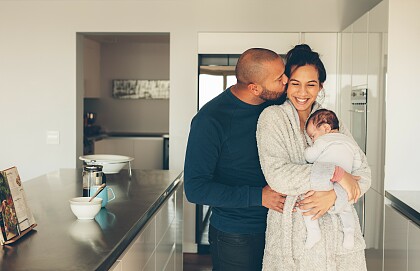 Eine Frau mit einem Baby auf dem Arm und ein Mann stehen in einer Küche. Er küsst sie.