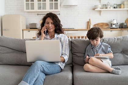 Frau sitzt telefonierend vor einem Laptop, rechts daneben ihr Sohn mit einem Tablet.